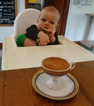 A baby in a high chair with an espresso coffee in front of him